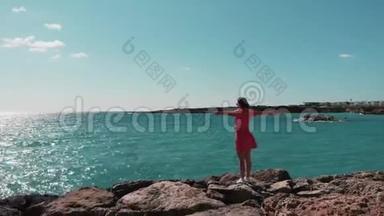 在阳光明媚的夏日，塞浦路斯珊瑚湾，穿着红色衣服的女士双手分开站在岩石海滩悬崖码头上，招展。 Ae